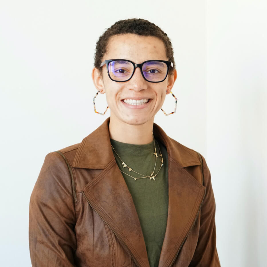 Emma with black rimmed glasses, a brown blazer, and green shirt.